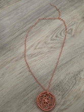 Load image into Gallery viewer, A delicate rose copper dreamcatcher necklace with a circular woven web pendant, laid out on a light gray wood-grain surface.