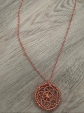 Load image into Gallery viewer, A rose gold metal necklace featuring a circular geometric pendant and a delicate chain, laid out on a gray wood-textured surface.