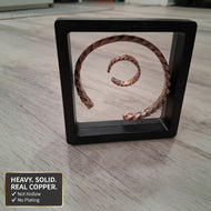 Copper spiral art piece in a black frame on a wooden surface