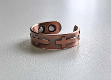 Load image into Gallery viewer, A copper-toned metal cuff bracelet with a cutout cross design and circular cutouts, resting on a light gray textured surface.
