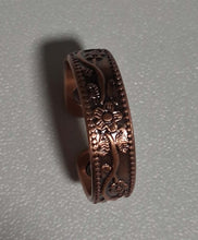 Load image into Gallery viewer, A copper-toned metal ring with an engraved floral pattern, resting on a textured gray surface.
