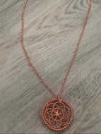 A rose gold metal necklace featuring a circular geometric pendant and a delicate chain, laid out on a gray wood-textured surface.