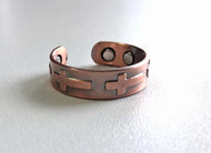 A copper-toned open cuff bracelet with a cross cutout design, resting on a white textured surface.