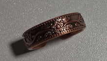 Load image into Gallery viewer, A copper-colored metal cuff bracelet with intricate floral engravings, resting on a textured gray surface.
