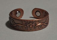 A copper-toned metal cuff bracelet with an embossed floral pattern and circular cutouts on the ends, displayed on a neutral gray background.